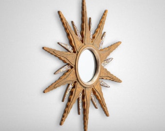 May include: A gold sunburst mirror with a round mirror in the center. The mirror has a wood frame with a textured surface and is decorated with small, iridescent glitter.