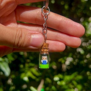 May include: A miniature glass bottle keychain with a cork stopper. Inside the bottle is a small figurine of a character wearing a blue shirt and a white hat, standing on a green base. The keychain has a silver ring and chain.