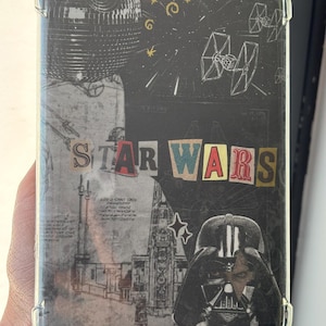 May include: A rectangular clear phone case featuring a Star Wars collage design. The graphic includes a Death Star, TIE fighters, a building structure, and Darth Vader. The text STAR WARS is printed in a multi-colored, cut-out letter style.
