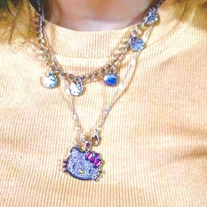 May include: A silver chain necklace with multiple charms featuring a Hello Kitty design. The charms are silver with a white enamel Hello Kitty face. The necklace also has a larger Hello Kitty charm with pink accents and rhinestones.