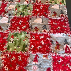 May include: A patchwork quilt featuring a festive Christmas theme. The quilt is composed of squares in red, green, and gray, adorned with gnomes, snowmen, and winter scenes. The edges of each square have a ruffled trim.