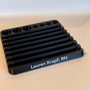 May include: Black plastic organizer with multiple slots for holding tools or other small items. The organizer has a personalized inscription that reads "Lauren Krapt, RN".