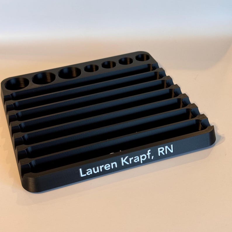 May include: Black plastic organizer with multiple slots for holding tools or other small items. The organizer has a personalized inscription that reads "Lauren Krapt, RN".