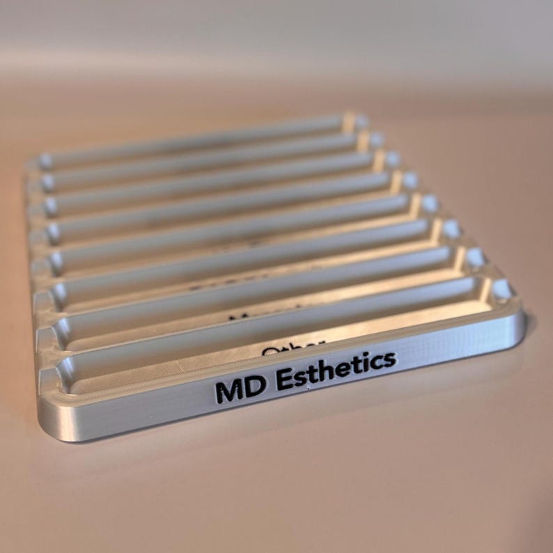 May include: A silver metal tray with seven slots for holding small items. The tray has the words "MD Esthetics" printed on the front.