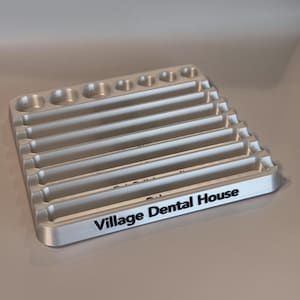 May include: A silver-colored tray with multiple rows of slots for holding dental tools. The tray has a black inscription that reads "Village Dental House".
