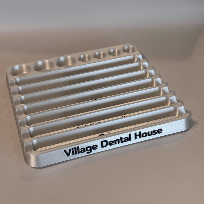 May include: A silver-colored tray with multiple rows of slots for holding dental tools. The tray has a black inscription that reads "Village Dental House".