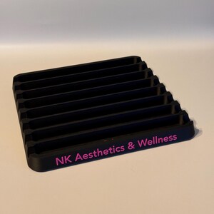 May include: Black plastic organizer with eight slots for storing items. The organizer has the text "NK Aesthetics & Wellness" printed in pink on the front.