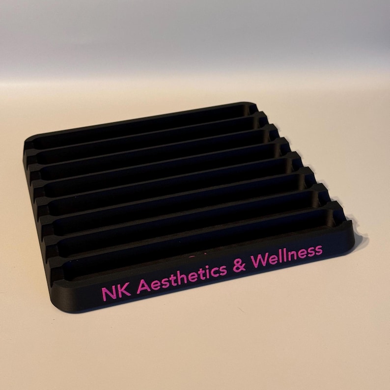 May include: Black plastic organizer with eight slots for storing items. The organizer has the text "NK Aesthetics & Wellness" printed in pink on the front.