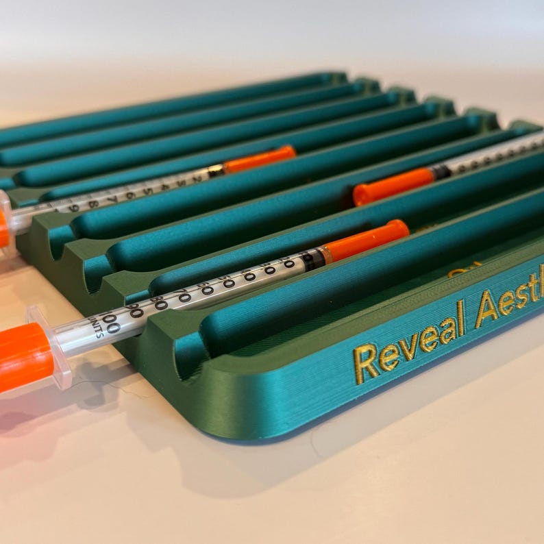 May include: Teal plastic organizer with six slots for syringes. The organizer has the text "Reveal Aesthetic" printed in gold on the front.