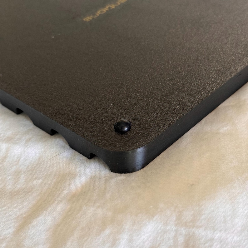May include: Close-up of a black plastic square with a small black rubber foot on the bottom corner. The square has a textured surface and the word "remarkable" is printed in gold on the top.