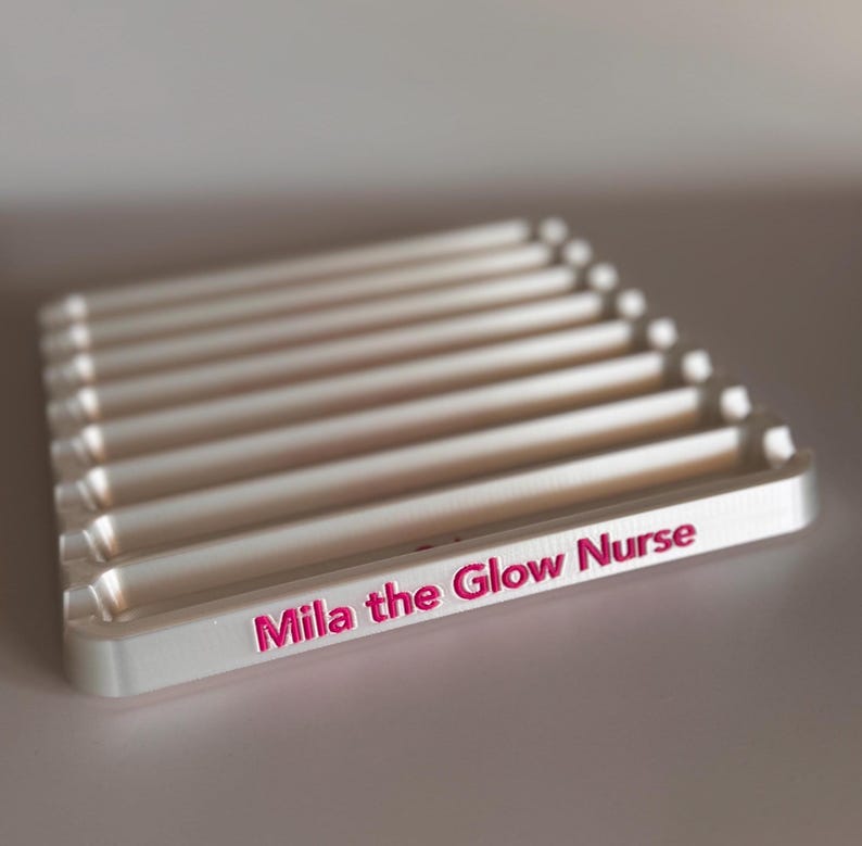 May include: White plastic tray with multiple slots for holding items. The tray has the words "Mila the Glow Nurse" printed in pink on the front.