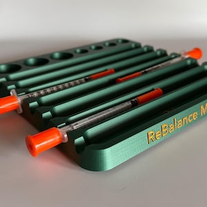 May include: A green plastic organizer with multiple slots for storing syringes. The organizer has the text "ReBalance" printed in gold on the side.