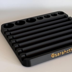 May include: Black plastic organizer with multiple slots and compartments for storing small items. The organizer has a textured surface and a gold inscription that reads "@sarahcth".