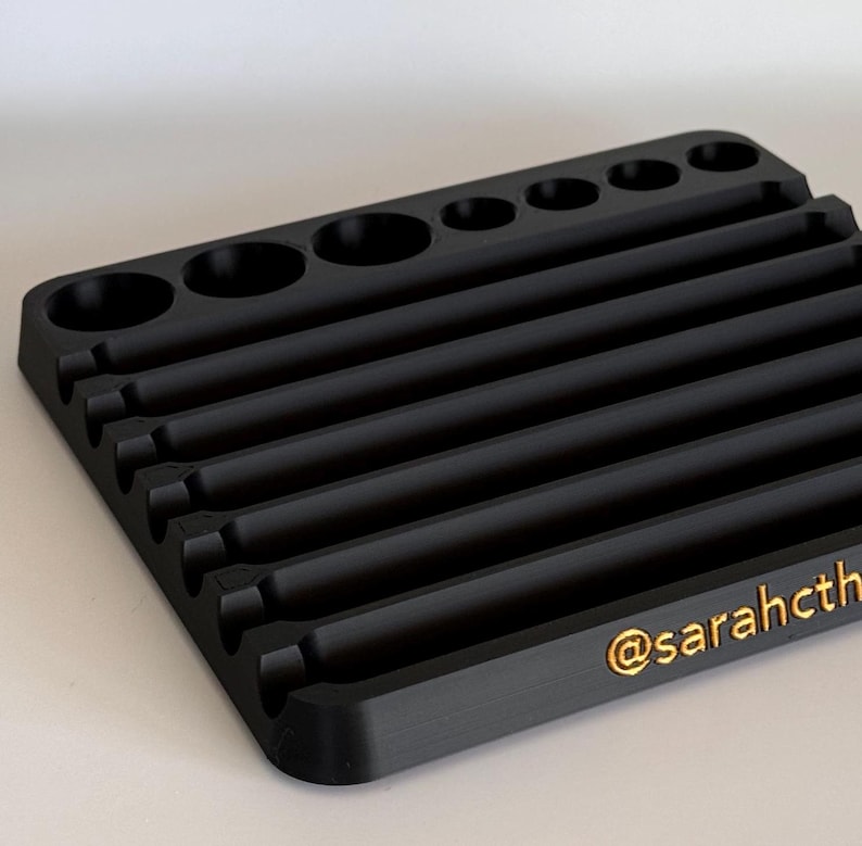 May include: Black plastic organizer with multiple slots and compartments for storing small items. The organizer has a textured surface and a gold inscription that reads "@sarahcth".