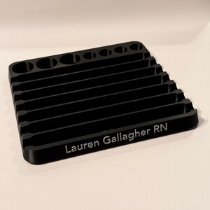 May include: Black plastic organizer with multiple slots and a rounded top. The organizer has the name "Lauren Gallagher RN" engraved on the front.