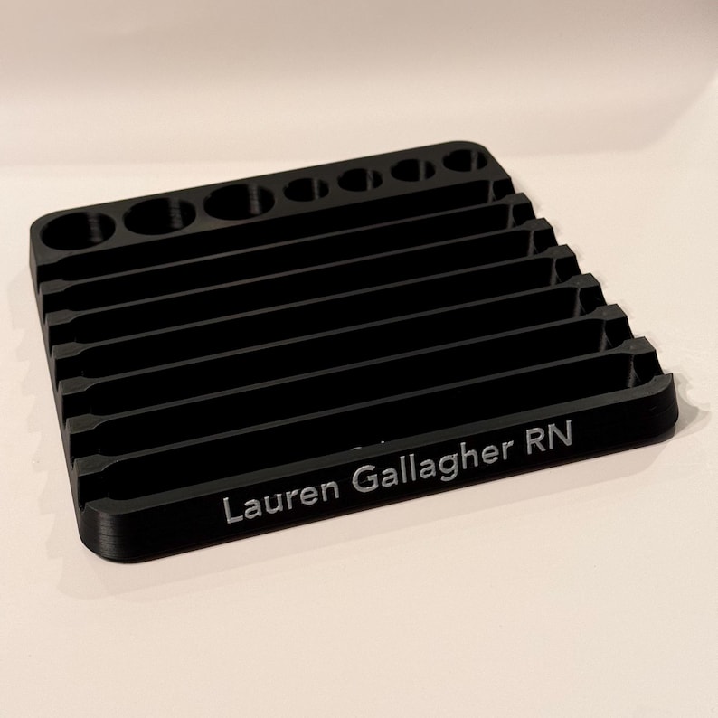 May include: Black plastic organizer with multiple slots and a rounded top. The organizer has the name "Lauren Gallagher RN" engraved on the front.