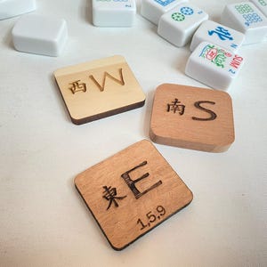 May include: Wooden Mahjong tile set with engraved Chinese characters and English letters. The tiles are square and brown, with the letters "W", "S", and "E" visible. Other white tiles are scattered around.