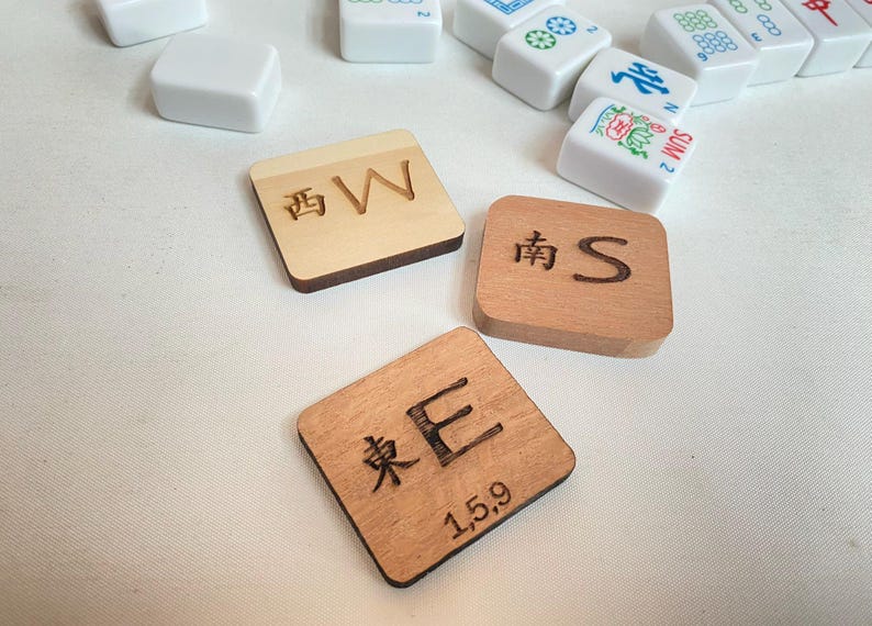 May include: Wooden Mahjong tile set with engraved Chinese characters and English letters. The tiles are square and brown, with the letters "W", "S", and "E" visible. Other white tiles are scattered around.
