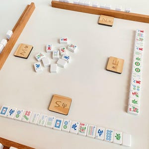 May include: A Mahjong game set with wooden racks and tiles. The tiles are white with colorful symbols and characters. Wooden tiles with the letters "M", "E", "N", and "S" are also present. The game is set on a white surface.