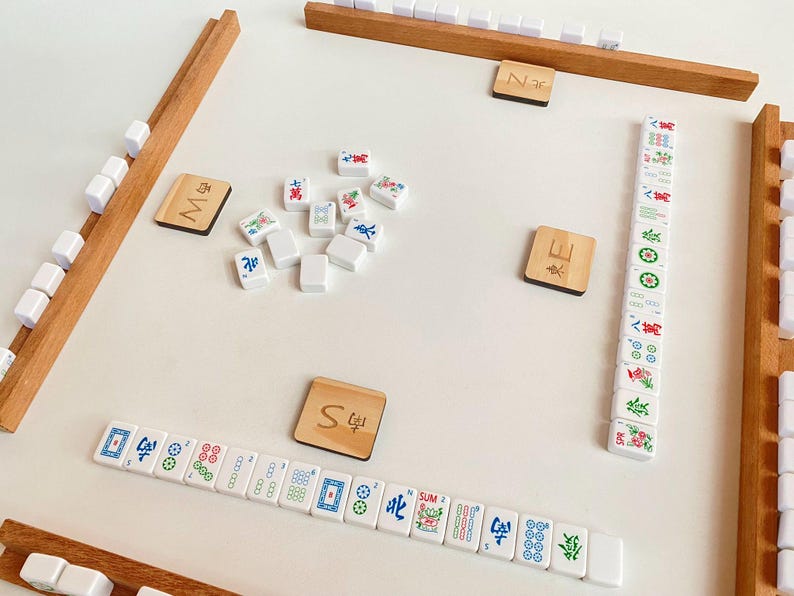May include: A Mahjong game set with wooden racks and tiles. The tiles are white with colorful symbols and characters. Wooden tiles with the letters "M", "E", "N", and "S" are also present. The game is set on a white surface.
