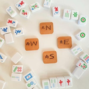 May include: A collection of white and colorful Mahjong tiles and four wooden direction tiles. The tiles feature Chinese characters and symbols in red, green, and blue. The wooden tiles are engraved with the cardinal directions.