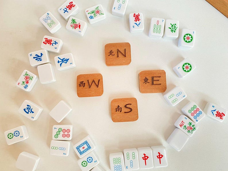 May include: A collection of white and colorful Mahjong tiles and four wooden direction tiles. The tiles feature Chinese characters and symbols in red, green, and blue. The wooden tiles are engraved with the cardinal directions.