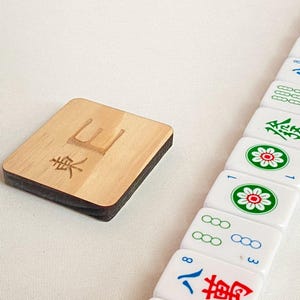 May include: A wooden Mahjong tile with the character "東" and a stack of white tiles with red, green, and blue symbols. The wooden tile is rectangular with a dark brown edge.