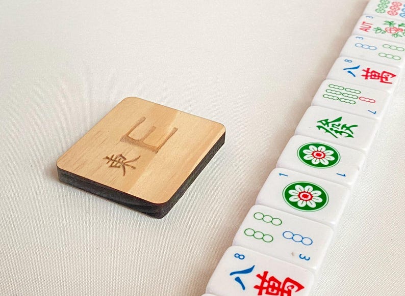 May include: A wooden Mahjong tile with the character "東" and a stack of white tiles with red, green, and blue symbols. The wooden tile is rectangular with a dark brown edge.