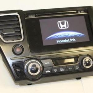 May include: A black Honda automotive dashboard infotainment unit featuring a central touchscreen display with the Honda logo and HondaLink text, integrated air vents, a hazard light button, and climate control knobs with silver trim accents.