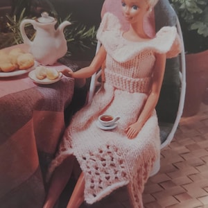 May include: A Barbie doll in a light pink knitted dress, seated at a table set for tea. A small white teapot, a plate of pastries, and a cup of tea are visible. The doll has blonde hair and is wearing white heels.
