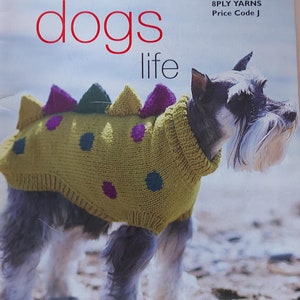 Può includere: Un piccolo cane bianco che indossa un maglione di lana verde con pois viola e blu. Il maglione ha un design a forma di dinosauro con punte sulla schiena. Il cane è in piedi su una spiaggia sabbiosa. L'immagine è tratta da un libro di modelli di maglia intitolato "It's a dogs life" di Patons.