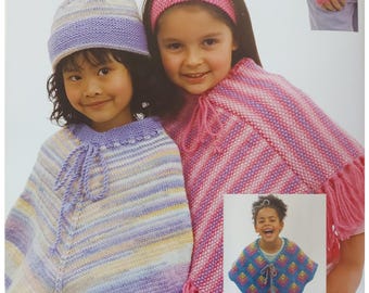 May include: Two girls wearing colorful striped knitted ponchos. One girl is wearing a pink headband and the other girl is wearing a purple and white beanie. The girls are smiling and looking at the camera.