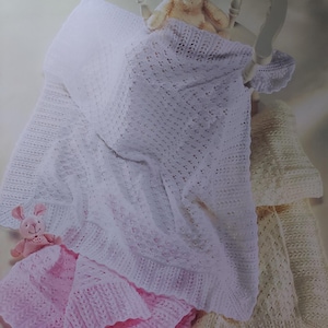 Könnte beinhalten: Drei gestrickte Babydecken in Weiß, Rosa und Creme. Die Decken sind mit einem Spitzenmuster gestrickt und werden auf einem weißen Stuhl mit einem Teddybär präsentiert.