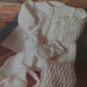 May include: A white knitted baby set comprising a cardigan, hat, booties, and a ribbon. The cardigan features a button fastening and a delicate lace design. The set is made with four-ply yarn and is 41-51cm in size.