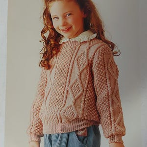 Peut inclure: Un pull rose en tricot avec un motif en câble. Le pull a un col rond et des manches longues.