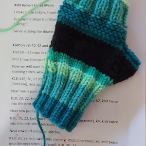 May include: A close-up of a partially knitted fingerless glove with a green, blue, and black striped pattern. The knitting instructions are visible in the background, with instructions to cast on 38, 40, or 42 stitches and knit 18-26 rows in knit.