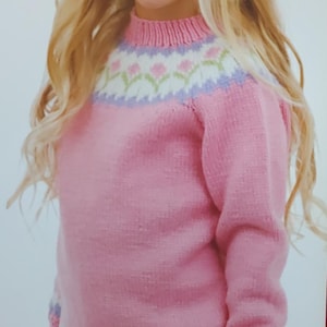 May include: A young girl wearing a pink sweater with a white and purple Fair Isle yoke design. The yoke features a row of small flowers. The sweater has a white trim around the neckline and cuffs. The girl is also wearing a white skirt.