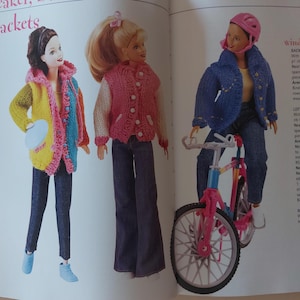 May include: Three dolls wearing colorful knitted jackets. The doll on the left is holding a blue frisbee. The doll in the middle is wearing a pink jacket with white sleeves. The doll on the right is wearing a blue jacket and riding a pink bicycle.