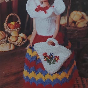 Puede incluir: Una muñeca con un top blanco de ganchillo con un lazo rojo, una falda a rayas rojas, amarillas y azules y un bolso blanco de ganchillo con flores bordadas.