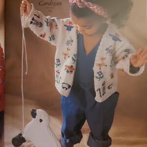 Pode incluir: Um cardigan branco de malha com um design de animais de fazenda. O cardigan é usado com uma faixa de cabeça xadrez vermelha, branca e azul. A criança está usando um macacão azul e brincando com um carneiro de brinquedo branco.