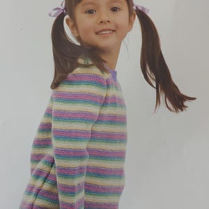 May include: A young girl wearing a multi-colored striped sweater with long sleeves. The sweater has stripes of pink, yellow, green, and blue. The girl has her hair pulled back in pigtails with purple bows.