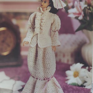May include: A crocheted doll outfit in white with a pink ribbon and a matching white crocheted hat. The doll is wearing a long dress with a jacket and a hat.