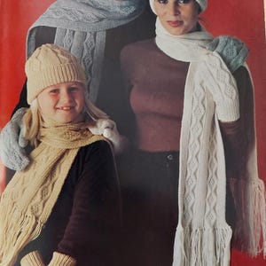 Könnte beinhalten: Eine Familie trägt passende Strickmützen und Schals. Die Mützen sind im Zopfmuster gestrickt und haben einen Bommel. Die Schals sind ebenfalls im Zopfmuster gestrickt und haben Fransen. Die Familie trägt braune, weiße und graue Kleidung.