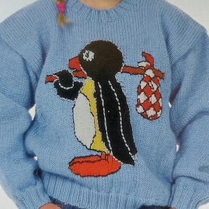 Puede incluir: Un suéter de punto azul claro con un diseño de pingüino. El pingüino lleva un gorro rojo y lleva una bolsa de cuadros rojos y blancos.