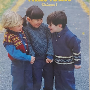 Puede incluir: Una portada de libro con tres niños que llevan suéteres tejidos. El libro se titula "Kids Knits" y es el volumen 3. La portada es una foto de los niños de pie en un camino con hojas de otoño.