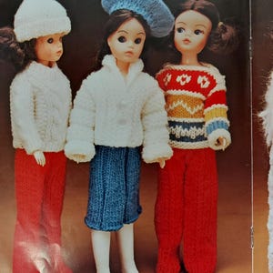 Könnte beinhalten: Drei Puppen, die gestrickte Kleidung tragen. Die Puppe links trägt einen weißen Hut, einen weißen Pullover und eine rote Hose. Die Puppe in der Mitte trägt eine blaue Baskenmütze, einen weißen Pullover, einen blauen Rock und weiße Stiefel. Die Puppe rechts trägt einen rot-gelb gestreiften Pullover mit rotem Blumenmuster und eine rote Hose.