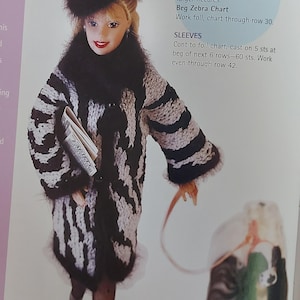 May include: A doll wearing a black and white zebra-striped coat with a matching hat and black fur trim. The doll is holding a small book and a leash attached to a small object.
