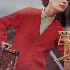 May include: A woman wearing a red cardigan with buttons, a white collared shirt, and a red beret. She is holding a brown suitcase and standing in front of a wooden structure.