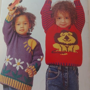 May include: Two children wearing knitted sweaters. The girl on the left is wearing a purple sweater with a daisy and sun design. The girl on the right is wearing a red sweater with a yellow dog design. Both girls are wearing blue jeans.