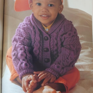 May include: A child wearing a purple cable-knit cardigan with three dark buttons, a yellow shirt, and orange pants. The child is also wearing an orange baseball cap. The child is sitting down and smiling.